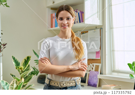 Portrait of smiling female teenager looking at camera with crossed arms in home 117335792