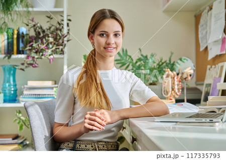 Portrait of smiling female teenager looking at camera, sitting at home at desk with computer Portrait of smiling female teenager looking at camera, sitting at home at desk with computer 117335793