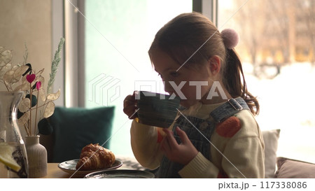 A girl drinks hot cocoa with marshmallows from a green cup against the backdrop of a cozy cafe with large panoramic windows. A girl drinks hot cocoa with marshmallows from a green cup against the backdrop of a cozy cafe with large panoramic windows. 117338086