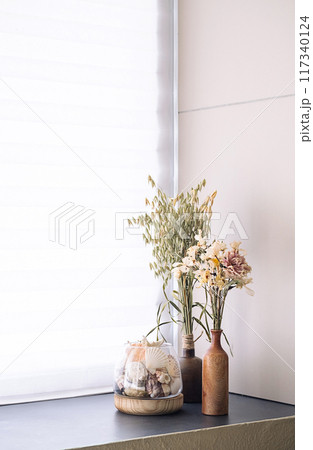Dried floral arrangement and seashells in rustic vases on windowsill by window. 117340124
