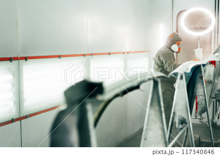 Auto mechanic worker wearing protective workwear spraying white paint on car part at workshop 117340846