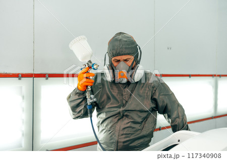 Auto mechanic worker wearing protective workwear spraying white paint on car part at workshop 117340908