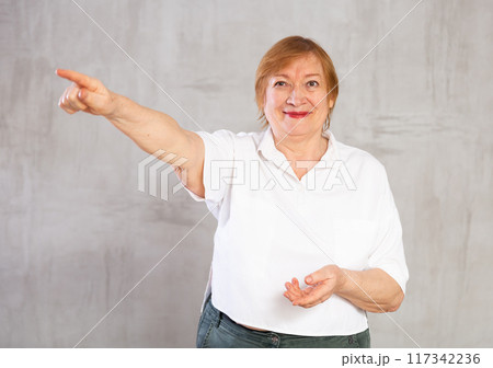 Smiling aging woman in white shirt gesturing towards with hand 117342236