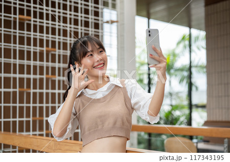 A charming Asian woman is taking selfies on her smartphone while standing in the building. A charming Asian woman is taking selfies on her smartphone while standing in the building. 117343195