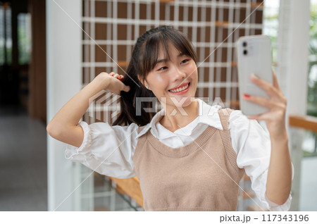 A charming Asian woman is taking selfies on her smartphone while standing in the building. 117343196