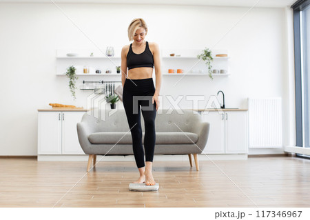 Woman in activewear checking weight on scale in modern living room Woman in activewear checking weight on scale in modern living room 117346967
