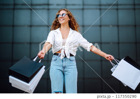Happy stylish woman with shopping bags enjoys sunny weather walking down street. Summer shopping. 117350290