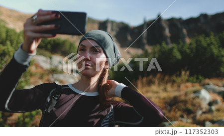 Active tourist woman holding cellphone to take selfie picture. Mountain nature landscape in background. Caucasian girl hiker enjoys moment. Travel, tourism, holiday, trekking, hiking, active lifestyle 117352303