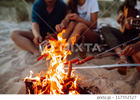 Fried sausages on bonfire. Group of young friends sitting on beach and fry sausages. Camping time. Fried sausages on bonfire. Group of young friends sitting on beach and fry sausages. Camping time. 117352527
