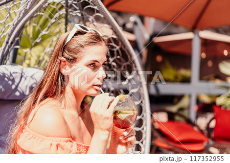 A woman in an orange dress is sitting in a chair with a drink in front of her. She is looking at the camera and she is enjoying her drink. The scene is set in a patio or outdoor area. A woman in an orange dress is sitting in a chair with a drink in front of her. She is looking at the camera and she is enjoying her drink. The scene is set in a patio or outdoor area. 117352955