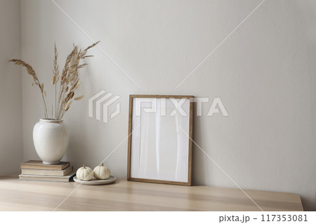 Elegant neutral poster mockup. Wooden picture frame on beige table. Vase with dry grass and old books and little white pumpkins. Empty wall background. Scandinavian interior, home. Autumn decor. 117353081