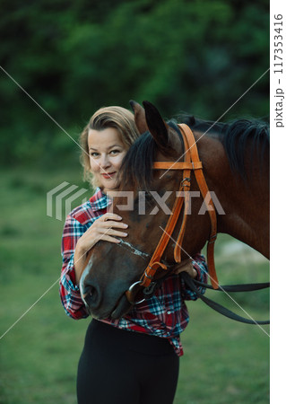 Happy blonde with horse in forest. Woman and a horse walking through the field during the day. Dressed in a plaid shirt and black leggings. 117353416