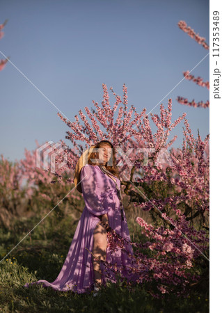Woman blooming peach orchard. Against the backdrop of a picturesque peach orchard, a woman in a long pink dress and hat enjoys a peaceful walk in the park, surrounded by the beauty of nature. 117353489