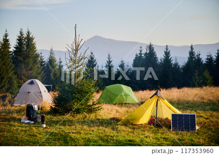 Solar panel in grassy field with tents in background highlights eco-friendly camping setup, surrounded by dense evergreen trees in evening. Integration of renewable energy in outdoor activities. 117353960