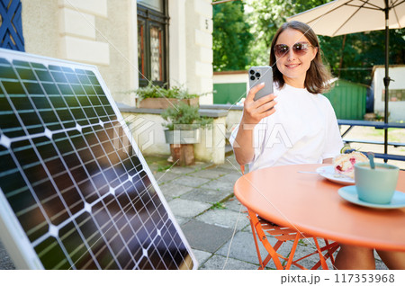 Young woman charging her smartphone from solar panel while relaxing. Pretty girl drinking coffee and checking her phone next to solar battery. Conscious female choosing sustainable lifestyle. 117353968