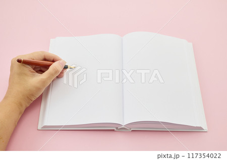 International Left-handers Day. Left hand holding a pen, writing in an open blank journal on a light pink background. 117354022