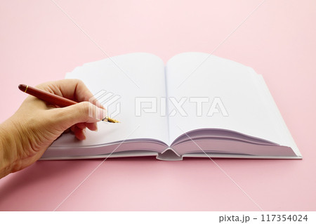 International Left-handers Day. Left Hand holding a pen, writing in an open blank journal on a light pink background. 117354024