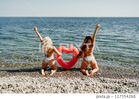 Two women are posing on a beach with a red heart-shaped inflatable raft 117354268