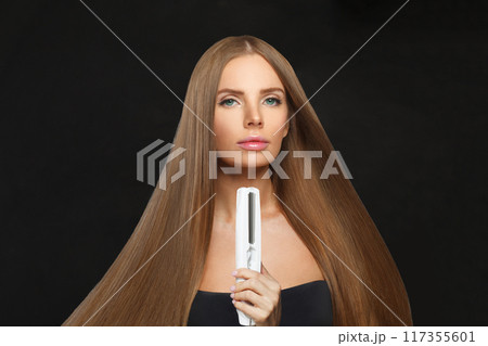 Charming woman with hair straightener iron in studio isolated on black background. Haircare 117355601