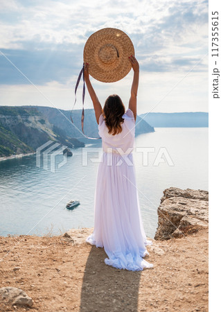 woman white dress stands on a rocky cliff overlooking the ocean. She is wearing a straw hat and she is enjoying the view. 117355615