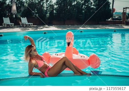 A woman is laying on a pink inflatable flamingo in a pool. The scene is bright and cheerful, with the pink flamingo adding a playful touch to the image. A woman is laying on a pink inflatable flamingo in a pool. The scene is bright and cheerful, with the pink flamingo adding a playful touch to the image. 117355617