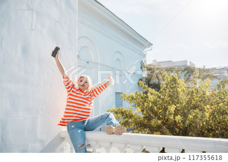 A woman is sitting on a ledge with her phone in her hand. She is wearing a striped shirt and blue jeans. The scene is set in a city with a tree in the background. 117355618