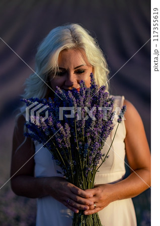 Blonde woman poses in lavender field at sunset. Happy woman in white dress holds lavender bouquet. Aromatherapy concept, lavender oil, photo session in lavender 117355619