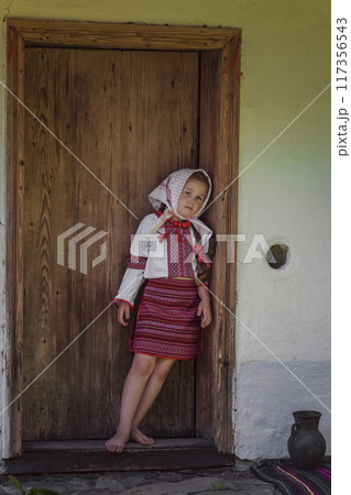 A delightful child wearing traditional dress stands joyfully by a rustic doorway A delightful child wearing traditional dress stands joyfully by a rustic doorway 117356543