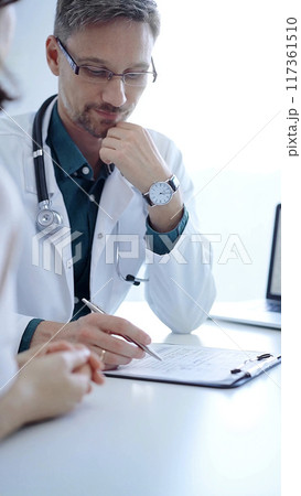 Doctor and a patient. The physician, wearing a white medical coat over a green shirt, gesturing with his hands during a consultation in the clinic. Medicine concept 117361510