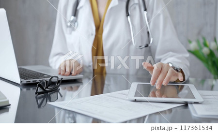 Doctor woman using tablet on the glass desk in medical office. Close-up of a doctor's hands interacting with a touch pad in clinic. Medicine and health care 117361534
