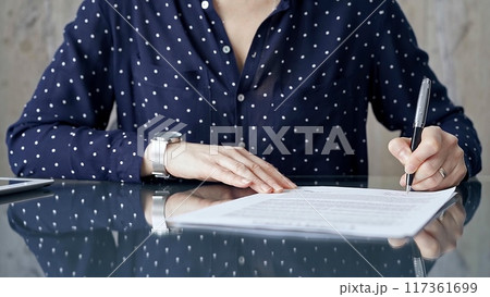 Businesswoman is signing corporate contract at her office desk in formal dark blue blouse with polka dots. Business people concept Businesswoman is signing corporate contract at her office desk in formal dark blue blouse with polka dots. Business people concept 117361699
