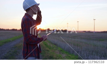 Man engineer wearing a white protective helmet is taking notes with a clipboard in a field with wind turbines, as the sun sets. Clean energy and engineering concept 117362110