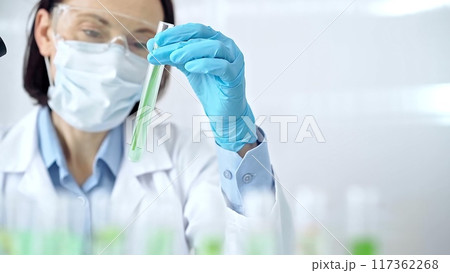 Woman scientist with protective gloves and glasses examining lab tube with a green fluid. Microbiology and science concept Woman scientist with protective gloves and glasses examining lab tube with a green fluid. Microbiology and science concept 117362268