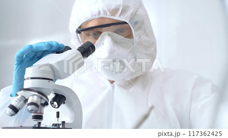 Scientist in ppe analyzing samples using a microscope in a laboratory setting. Close-up of a focused researcher in a lab coat mask and protective gear Scientist in ppe analyzing samples using a microscope in a laboratory setting. Close-up of a focused researcher in a lab coat mask and protective gear 117362425