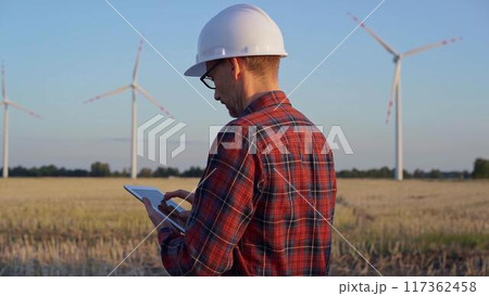 Man engineer at work is taking notes with a tablet computer in a field with wind turbines, as the sun sets. Clean energy concept Man engineer at work is taking notes with a tablet computer in a field with wind turbines, as the sun sets. Clean energy concept 117362458