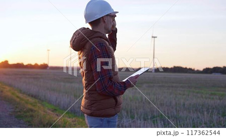 Man engineer, wearing a white protective helmet is taking notes with a tablet computer in a field with wind turbines, as the sun sets. Clean energy and engineering concept Man engineer, wearing a white protective helmet is taking notes with a tablet computer in a field with wind turbines, as the sun sets. Clean energy and engineering concept 117362544