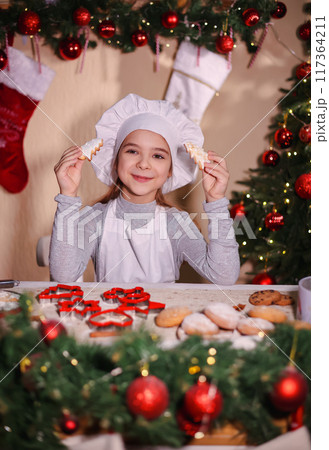 A cheerful girl in a chef's hat shows Christmas gingerbread cookies in the shape of a Christmas tree. 117364211