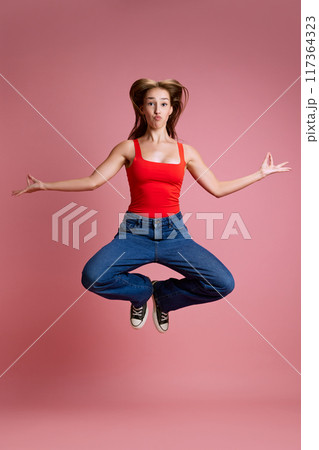 Full-length image of young beautiful girl in jeans and red top jumping in yoga pose against pink studio background. Meditation and energy Full-length image of young beautiful girl in jeans and red top jumping in yoga pose against pink studio background. Meditation and energy 117364323
