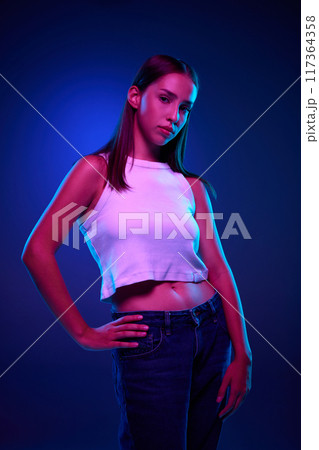 Portrait of beautiful young woman in casual clothes, white top and jeans posing with calm face against gradient blue studio background in neon light Portrait of beautiful young woman in casual clothes, white top and jeans posing with calm face against gradient blue studio background in neon light 117364358