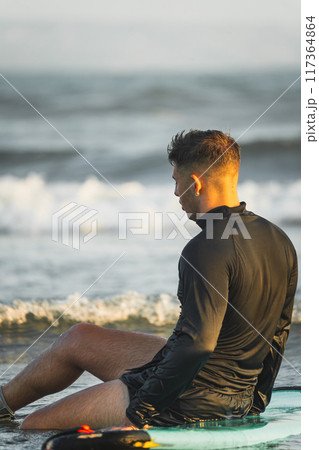 Surfer resting on his surfboard at the beach shore Surfer resting on his surfboard at the beach shore 117364864