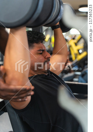 Young Latino man lifting weights at the gym with the help of his trainer 117365233