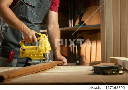 A skilled craftsman is focused on his woodworking project, using a electric jigsaw to cut timber precisely. The workshop is well-organized, with various tools and materials surrounding him, showcasing 117368806