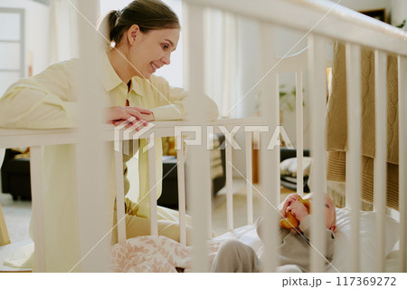 Smiling mother engaging with baby in crib holding a stuffed toy. Room decorated with warm, natural tones with calming atmosphere enhancing bond between parent and child Smiling mother engaging with baby in crib holding a stuffed toy. Room decorated with warm, natural tones with calming atmosphere enhancing bond between parent and child 117369272