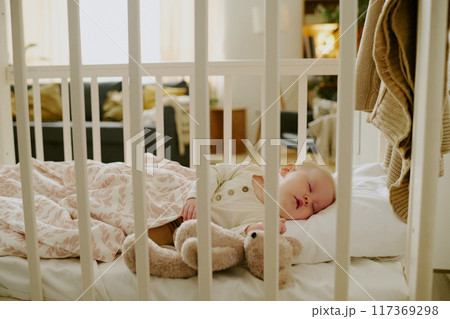 Baby sleeping peacefully in a crib, snuggling with a teddy bear, in a cozy room with natural light casting a soft glow Baby sleeping peacefully in a crib, snuggling with a teddy bear, in a cozy room with natural light casting a soft glow 117369298