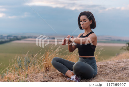 Woman is engaging in yoga outdoors with a fitness tracker, surrounded by scenic nature 117369407