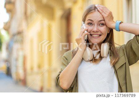 Happy surprised Caucasian young woman in pleasant conversation on smartphone standing on city street 117369789
