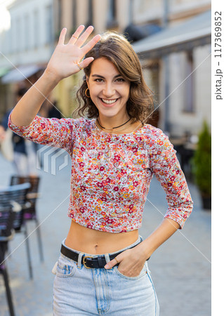 Young woman smiling friendly at camera, waving hands hello, hi, greeting or goodbye in city street 117369852