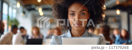 African American woman with afro hair holding a tablet while standing in front of people in the office. 117370268