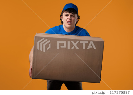 Postal courier man with heavy parcel box. Postman delivers address package on yellow studio background. 117370857