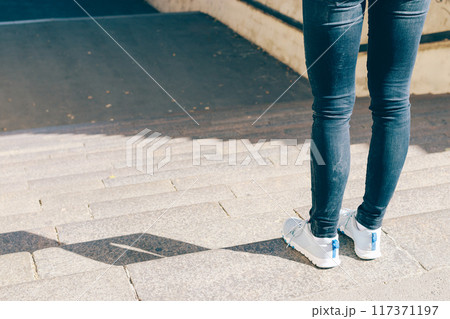 A woman wearing blue jeans and white shoes stands on a set of stairs 117371197